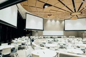 Image Title: Ready-to-Host Conference Room Image Description: Modern conference room with round tables, chairs, and large projector screens awaiting attendees Alt Text: Spacious event hall with set tables, chairs, and multiple projection screens Source: https://unsplash.com/photos/Q_KdjKxntH8