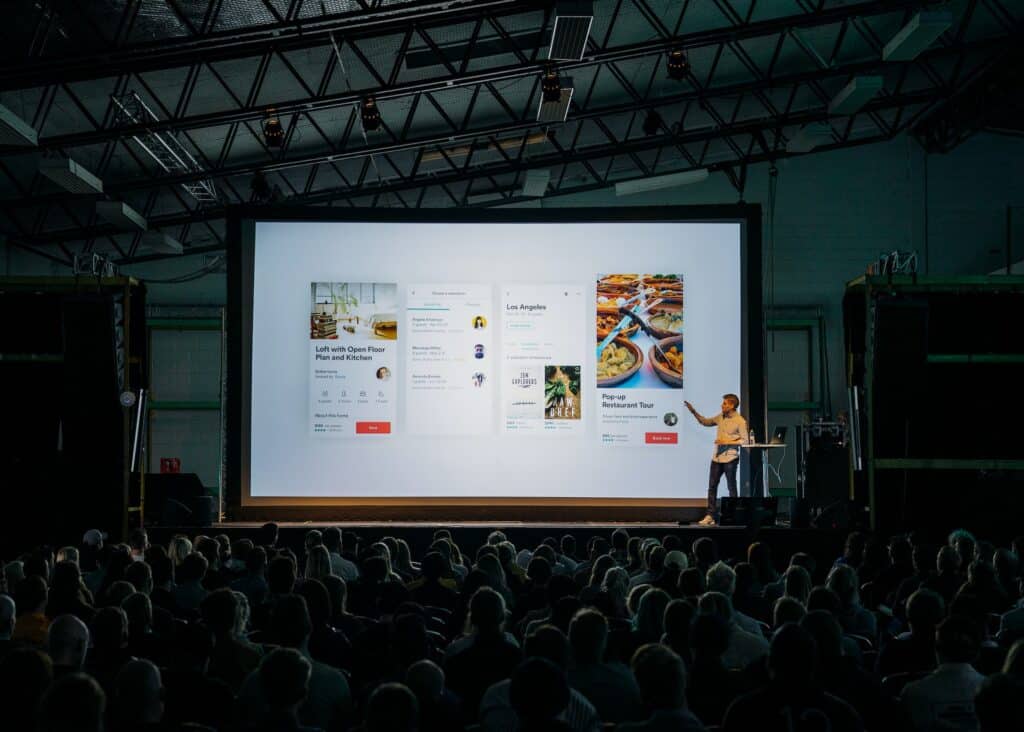 Image Title: Hybrid Event Spotlight Image Description: Presenter discussing a digital interface during a live event Alt Text: Event Presentation Source: https://unsplash.com/photos/person-discussing-while-standing-in-front-of-a-large-screen-in-front-of-people-inside-dim-lighted-room-bzdhc5b3Bxs