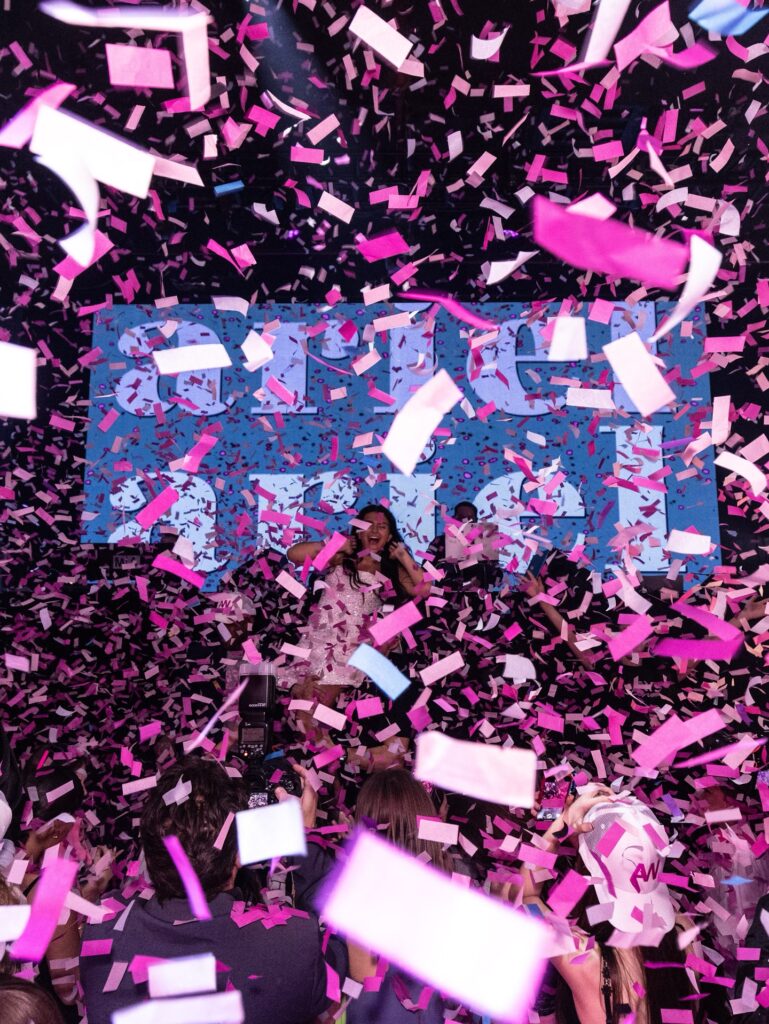 A young woman smiling on a stage as a large amount of pink and white confetti falls from the ceiling during a party. The crowd is cheering, and a large screen behind her displays a glowing logo