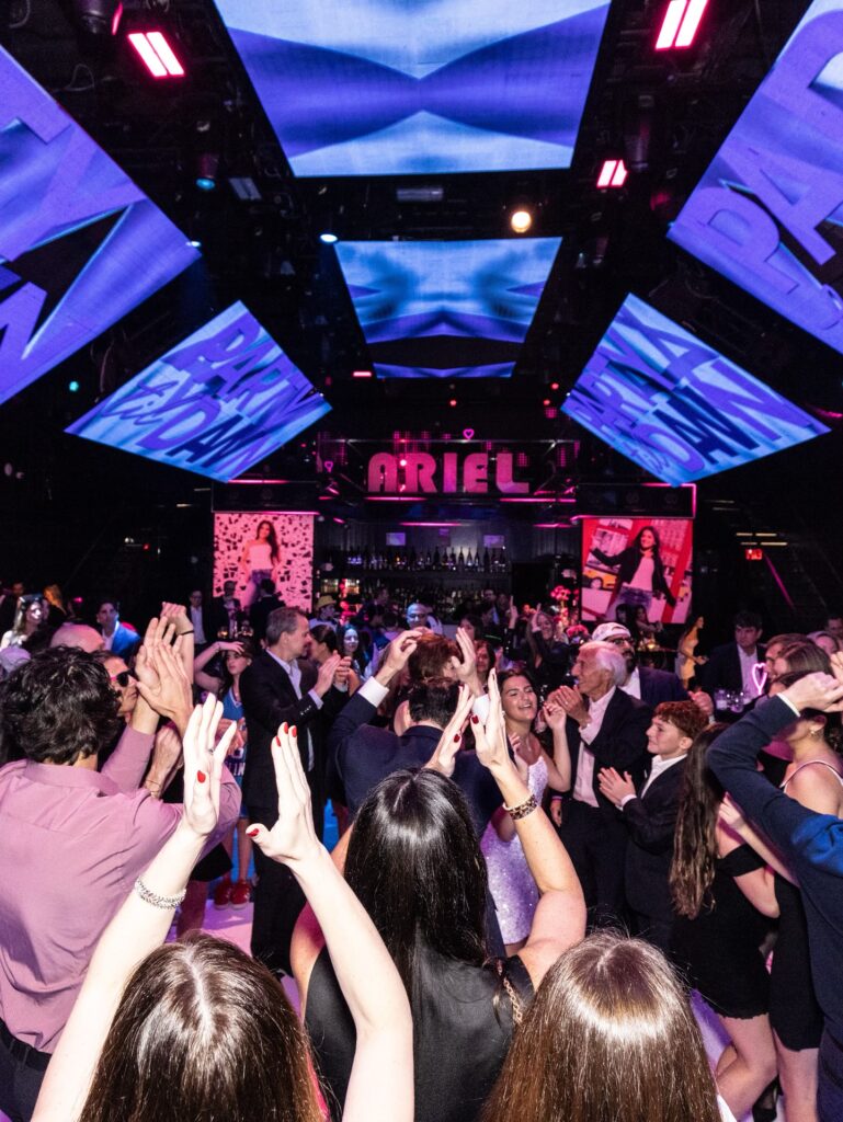 A vibrant party scene at a night event with a large crowd celebrating and dancing under blue and purple lights. The venue has a sign reading "ARIEL" and a large screen in the background.