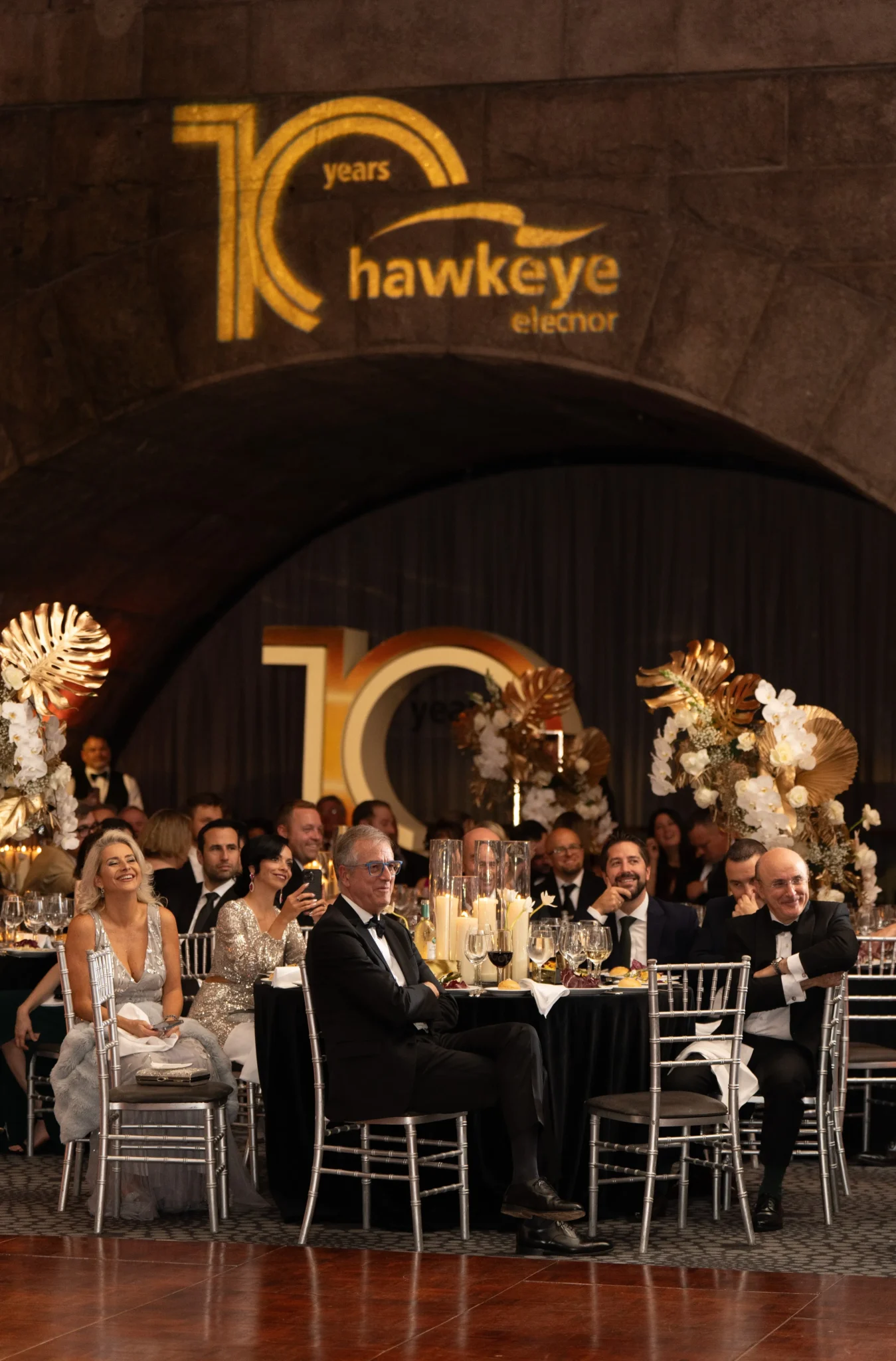 Guests seated at formal dinner tables beneath Hawkeye's illuminated 10th anniversary logo in NYC, EMRG Media