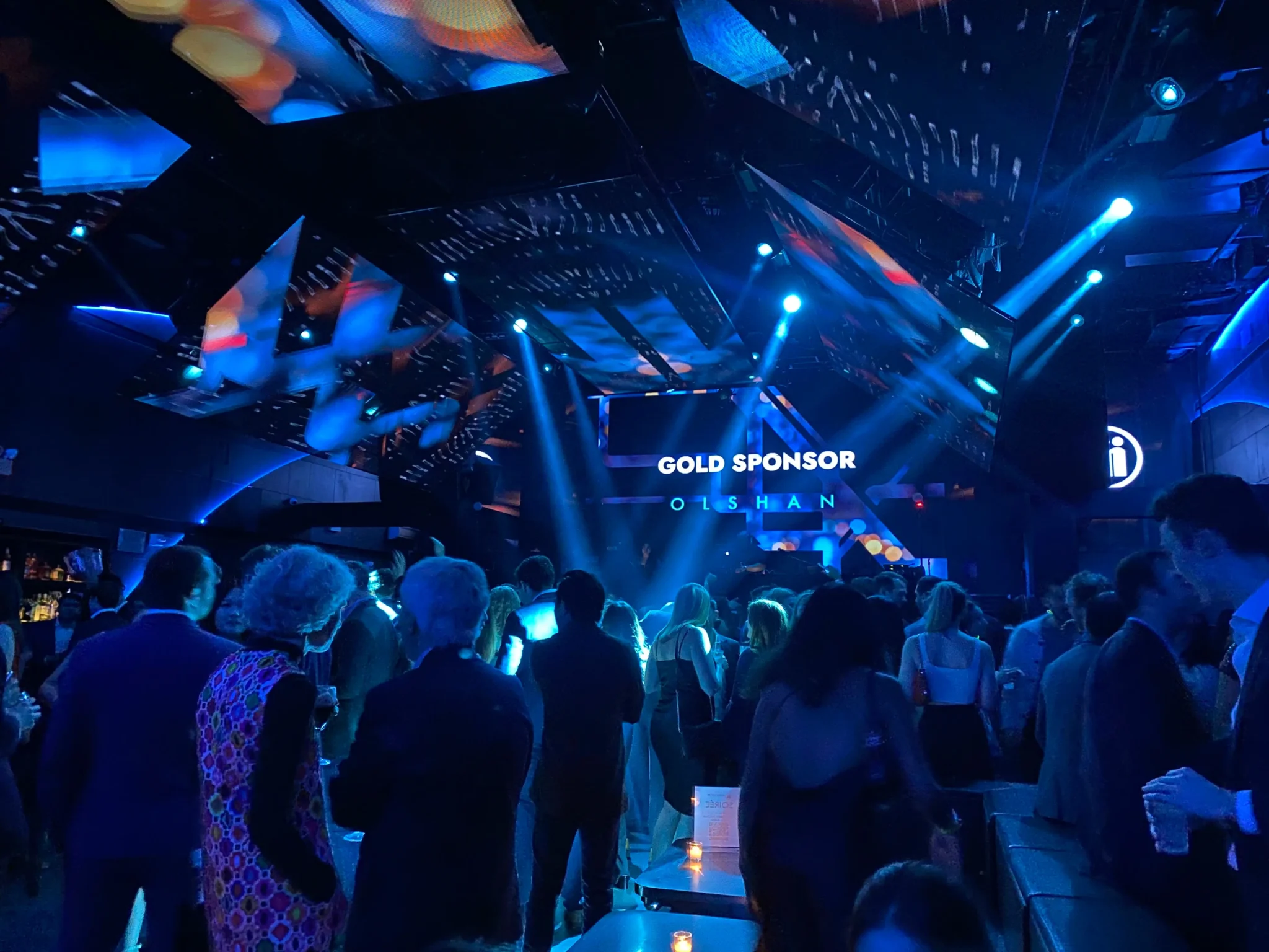 Crowd of guests under blue stage lighting with "Gold Sponsor Olshan" signage at a NYC event, EMRG Media